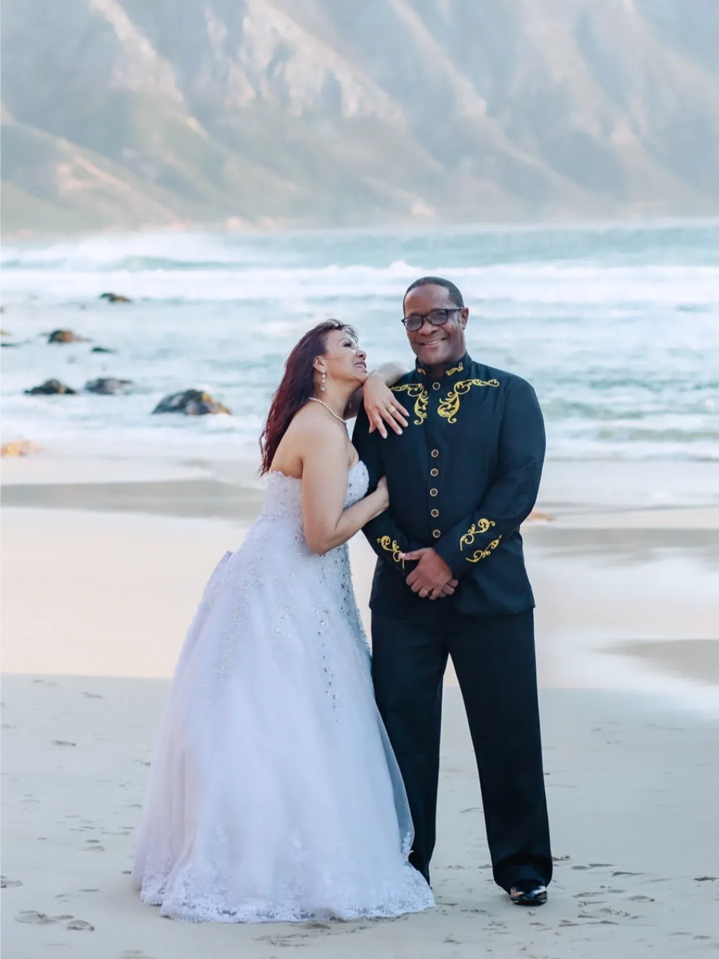 Bride leaning on her groom's shoulder on the beach, he in a black jacket with gold embroidery, surf and mountains behind.