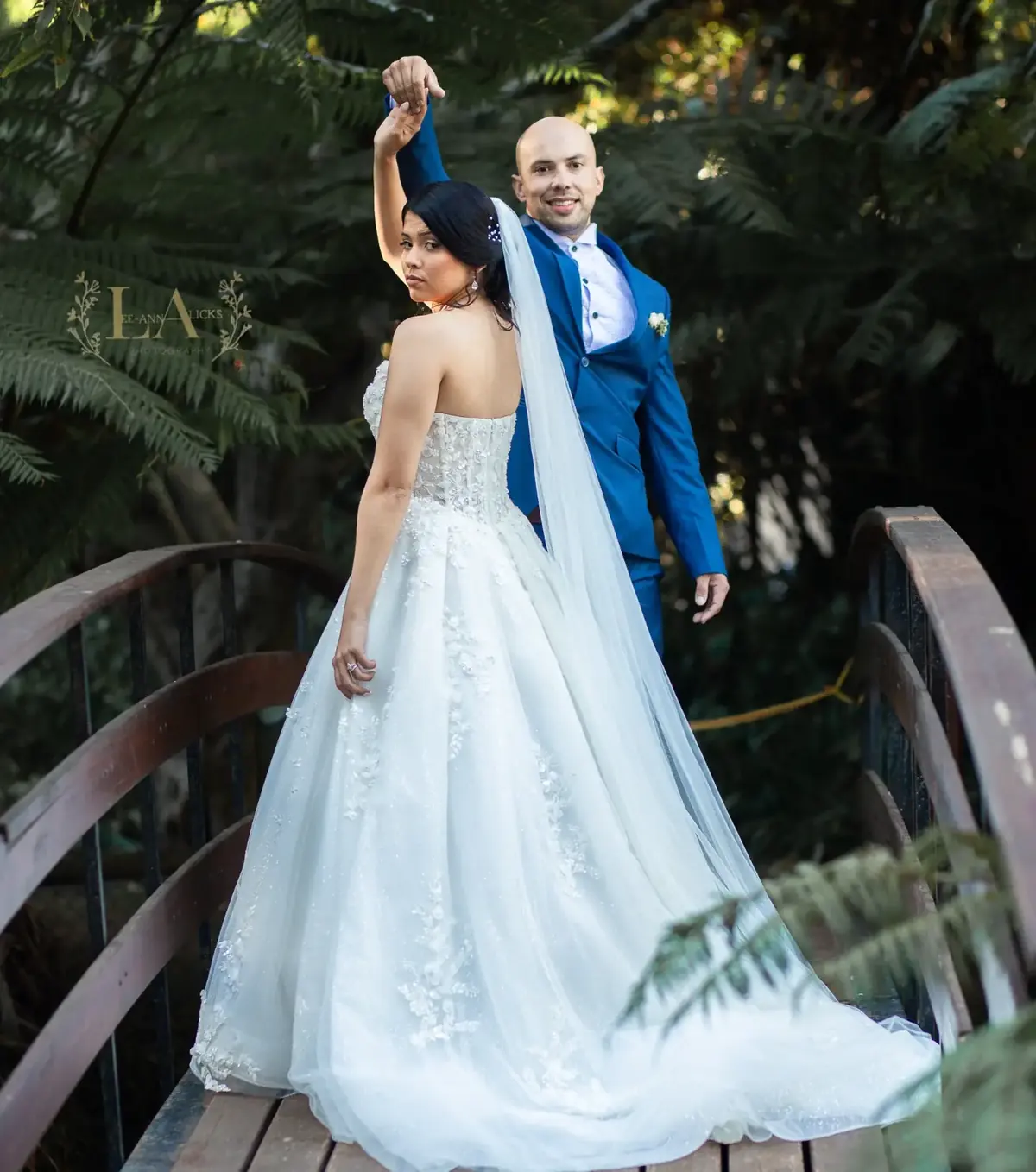 Groom twirling his bride on a wooden garden bridge, her veil and train trailing behind