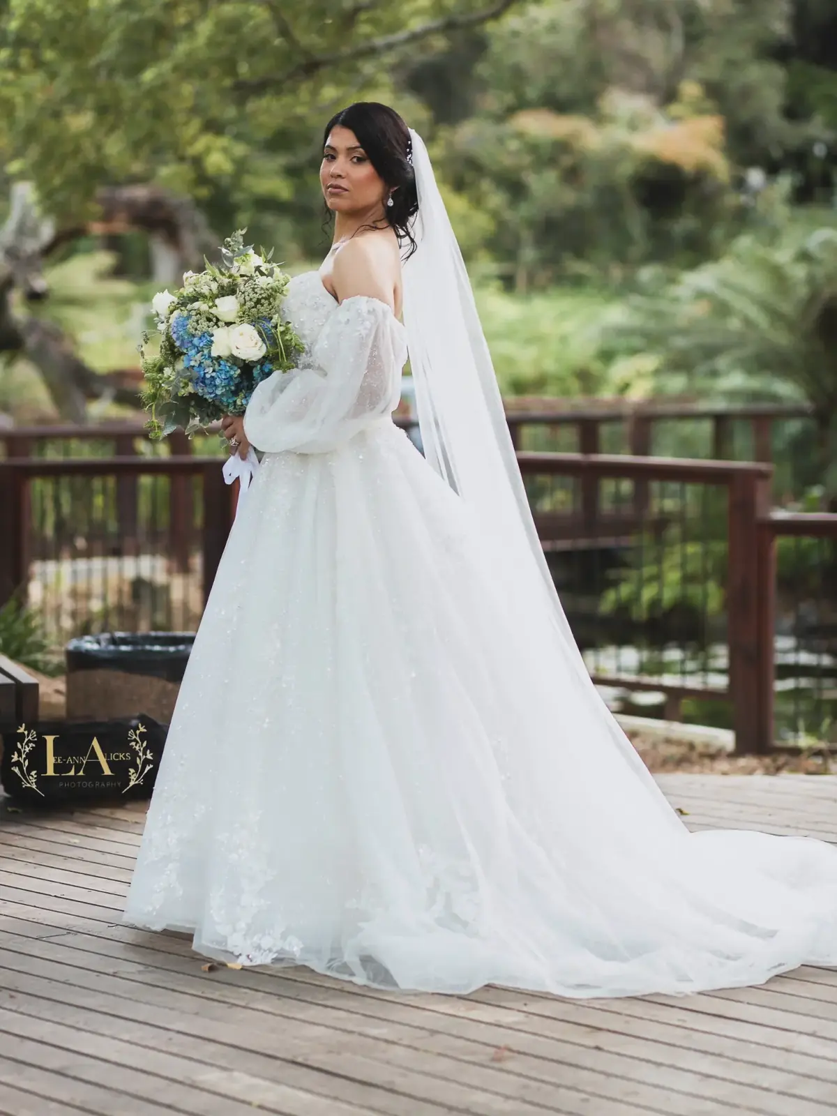 Bride in a lace gown with long sheer sleeves holding a blue and white bouquet on a garden deck