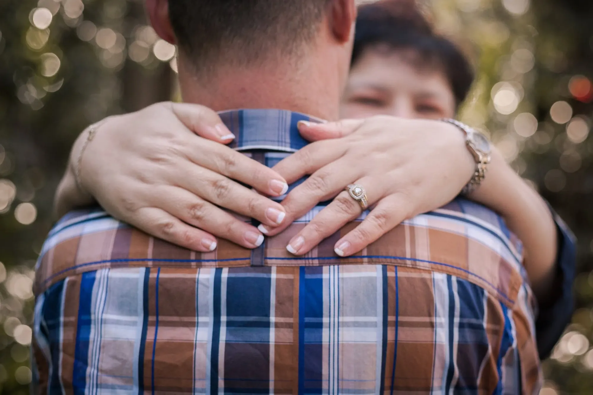 Close-up of her hands, with an engagement ring, crossed over his shoulders on a checked shirt during a couple shoot.