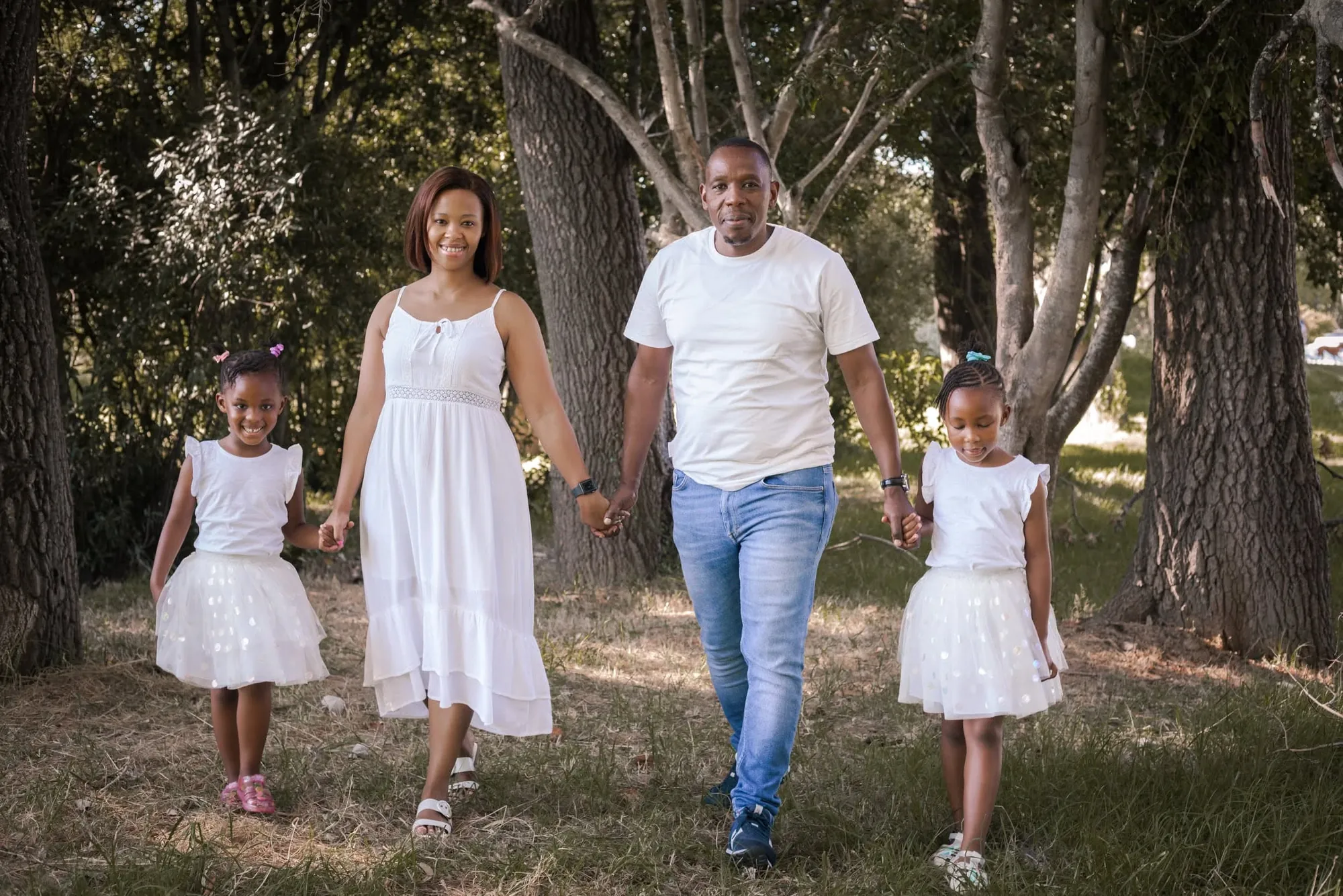 Family of four dressed in white walking hand in hand along a path under tall trees.
