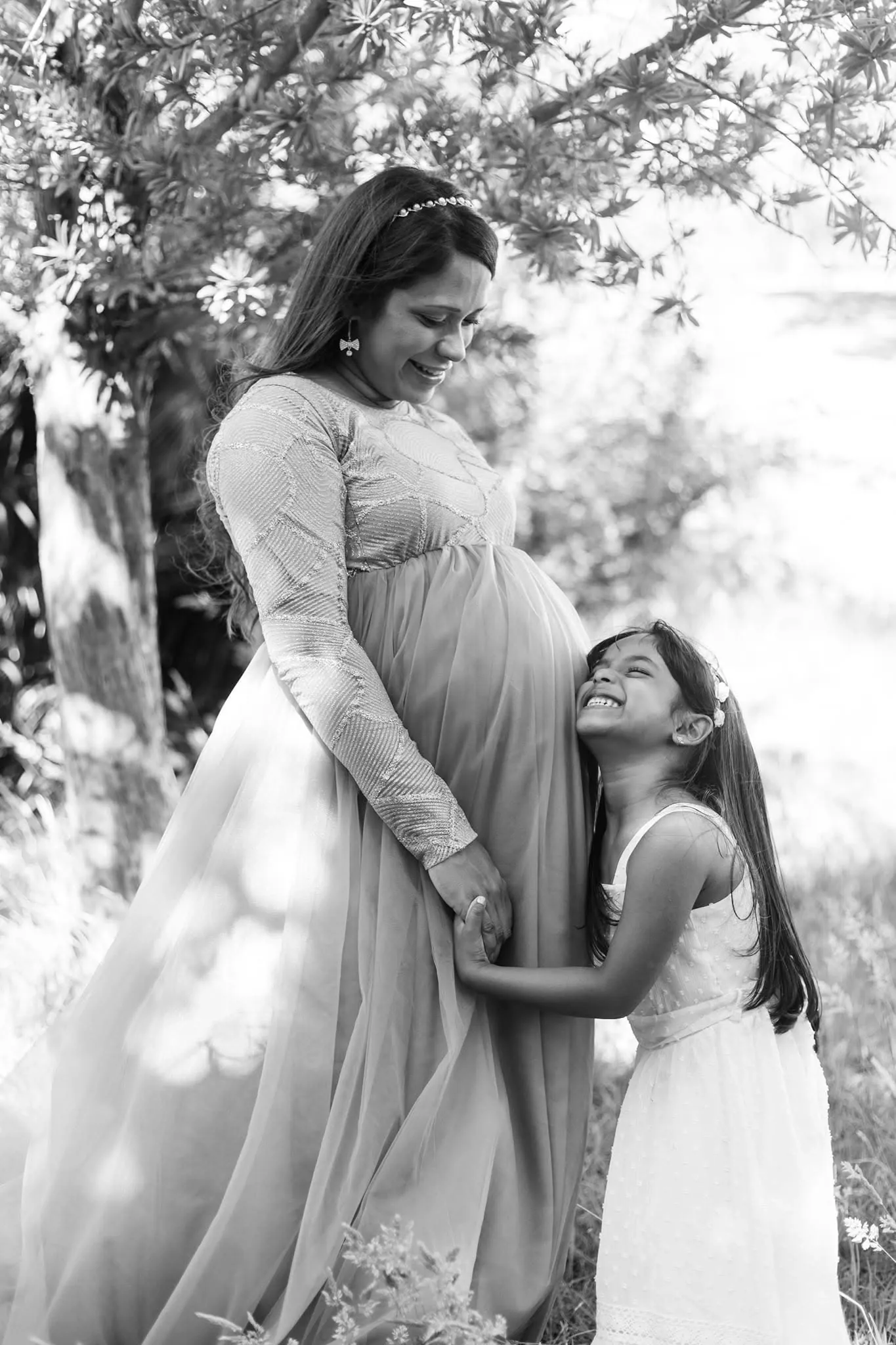 Black and white photo of a pregnant mum and her daughter, the girl resting her head on the bump.