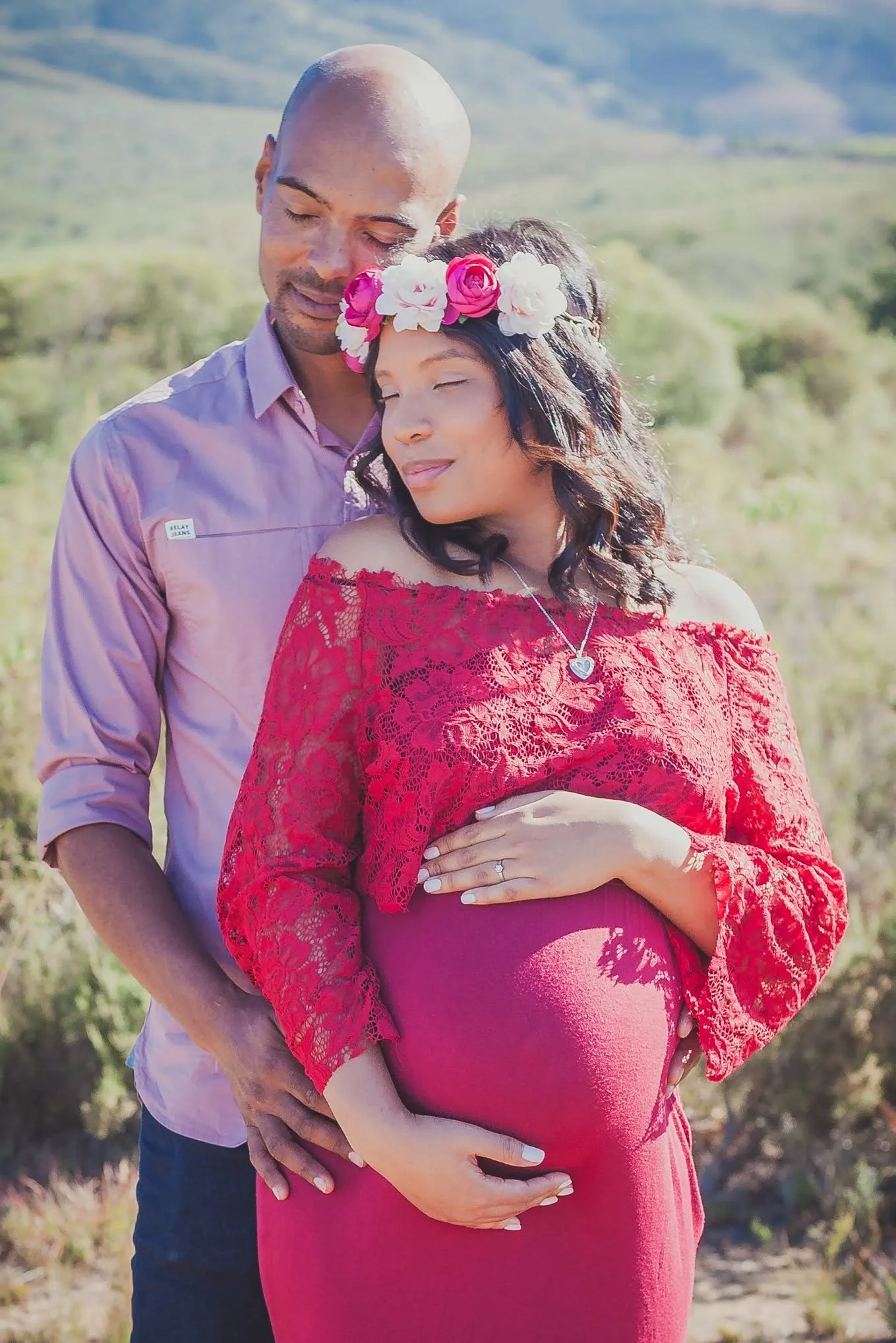 Couple at a maternity shoot, her in a red lace off-shoulder gown and flower crown, his hands on the bump.