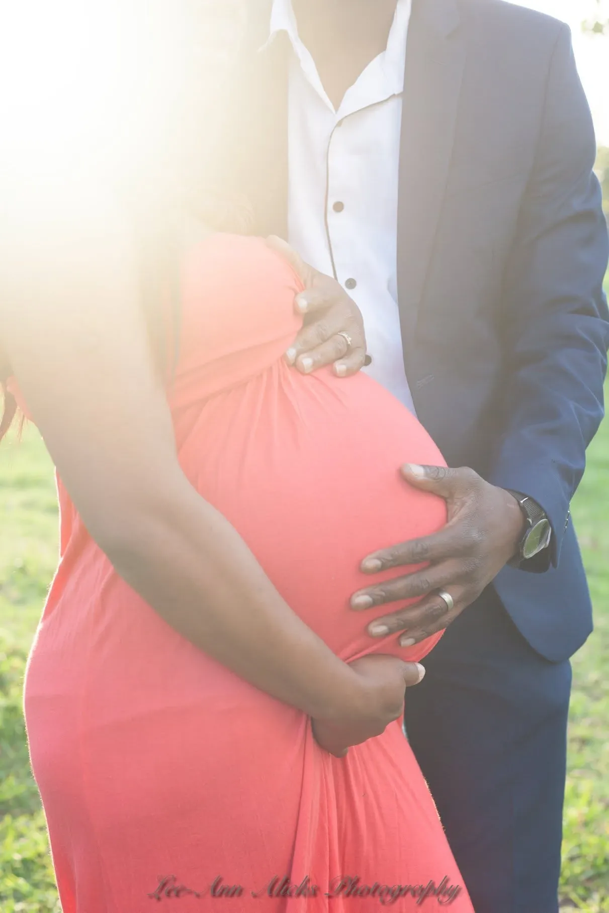Maternity photoshoot: partner's hands and hers on the bump, coral dress backlit by the late sun.