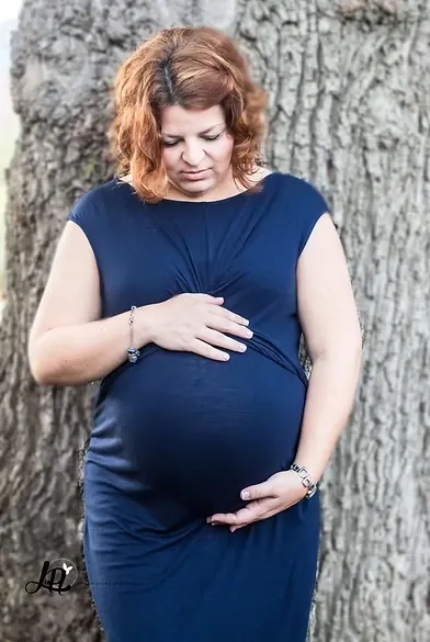 Mum-to-be in a navy dress looking down at her bump, hands resting on it, against old tree bark.