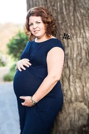 Mum-to-be in a navy dress leaning against a large tree trunk, smiling at the camera.