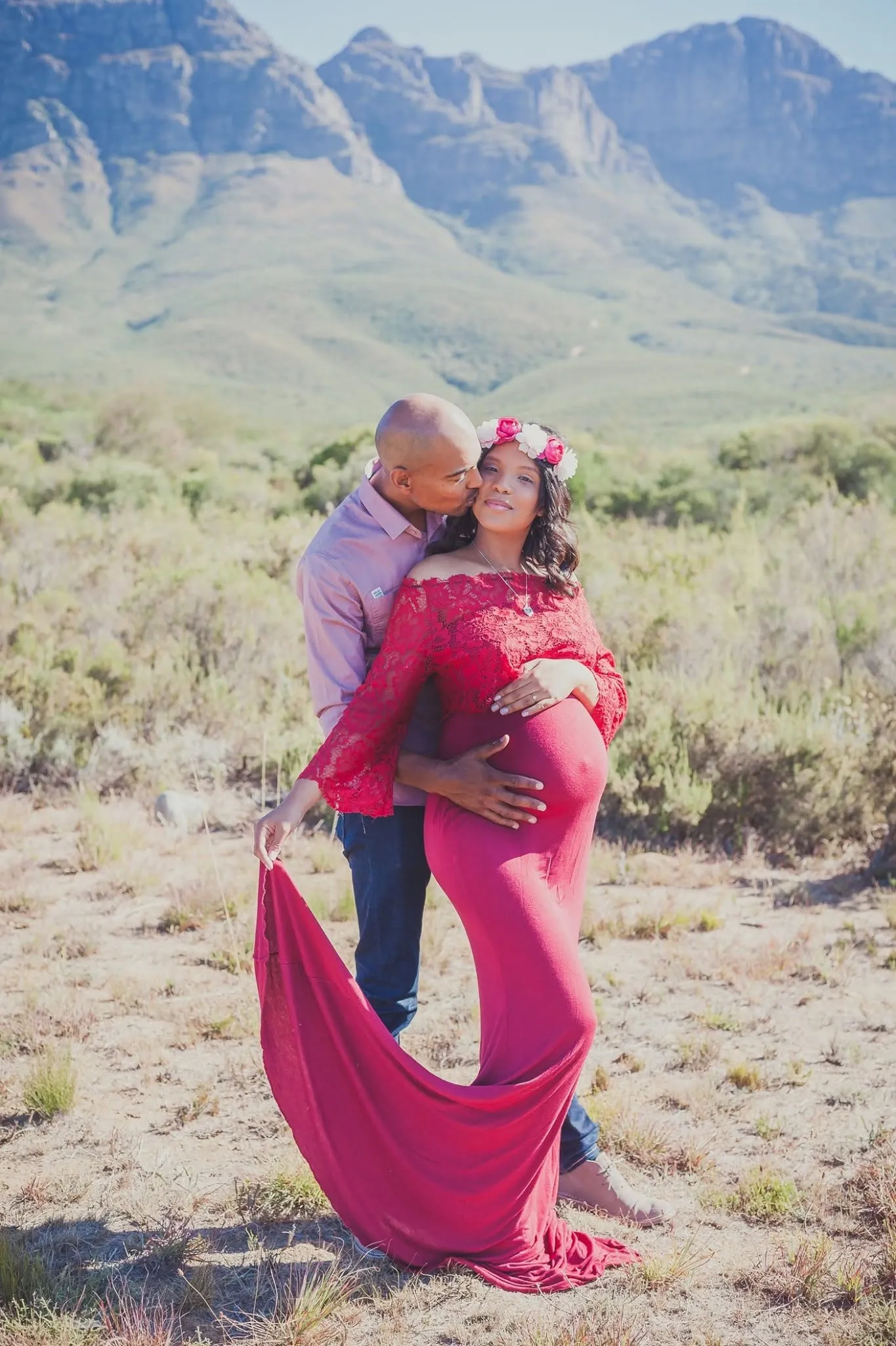 Pregnant woman in a red gown with a long train beside her partner in open veld, mountains behind.