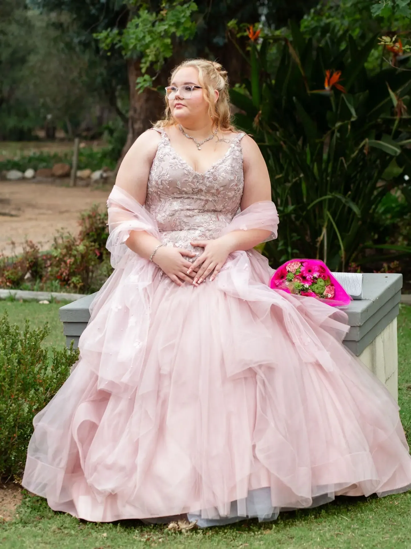 Matriculant seated on a garden bench in a pink gown with a bouquet beside her.