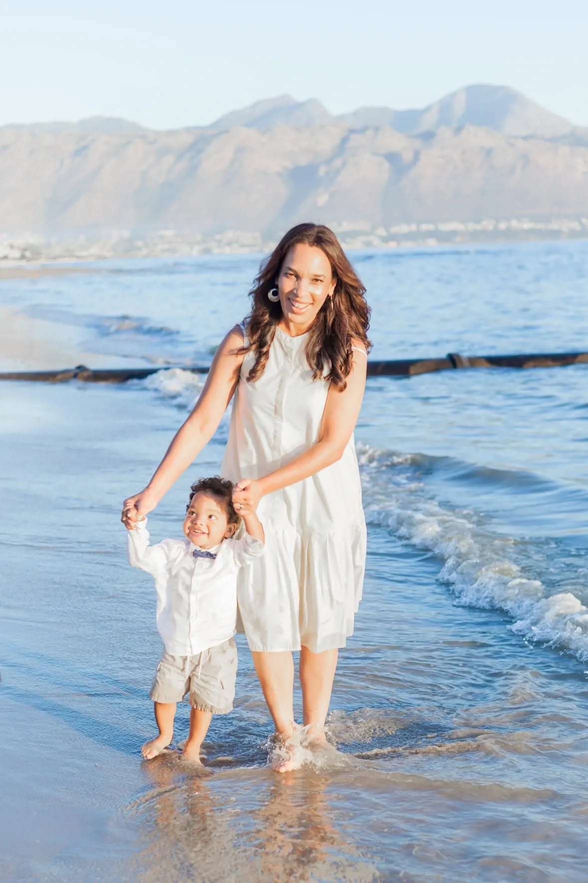 Mother holding her toddler's hands as he paddles in the shallows, mountains across the bay.