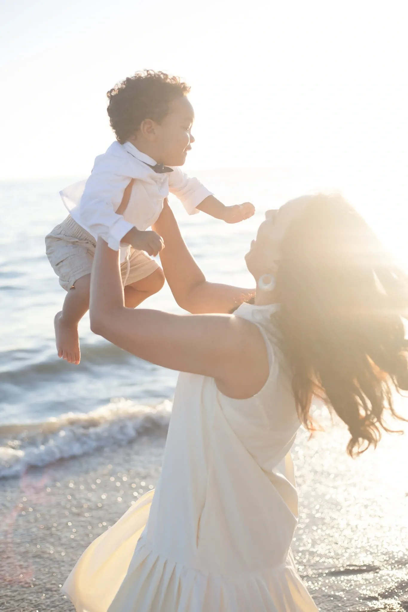 Mother lifting her toddler son into the air on the beach, backlit by the low sun.