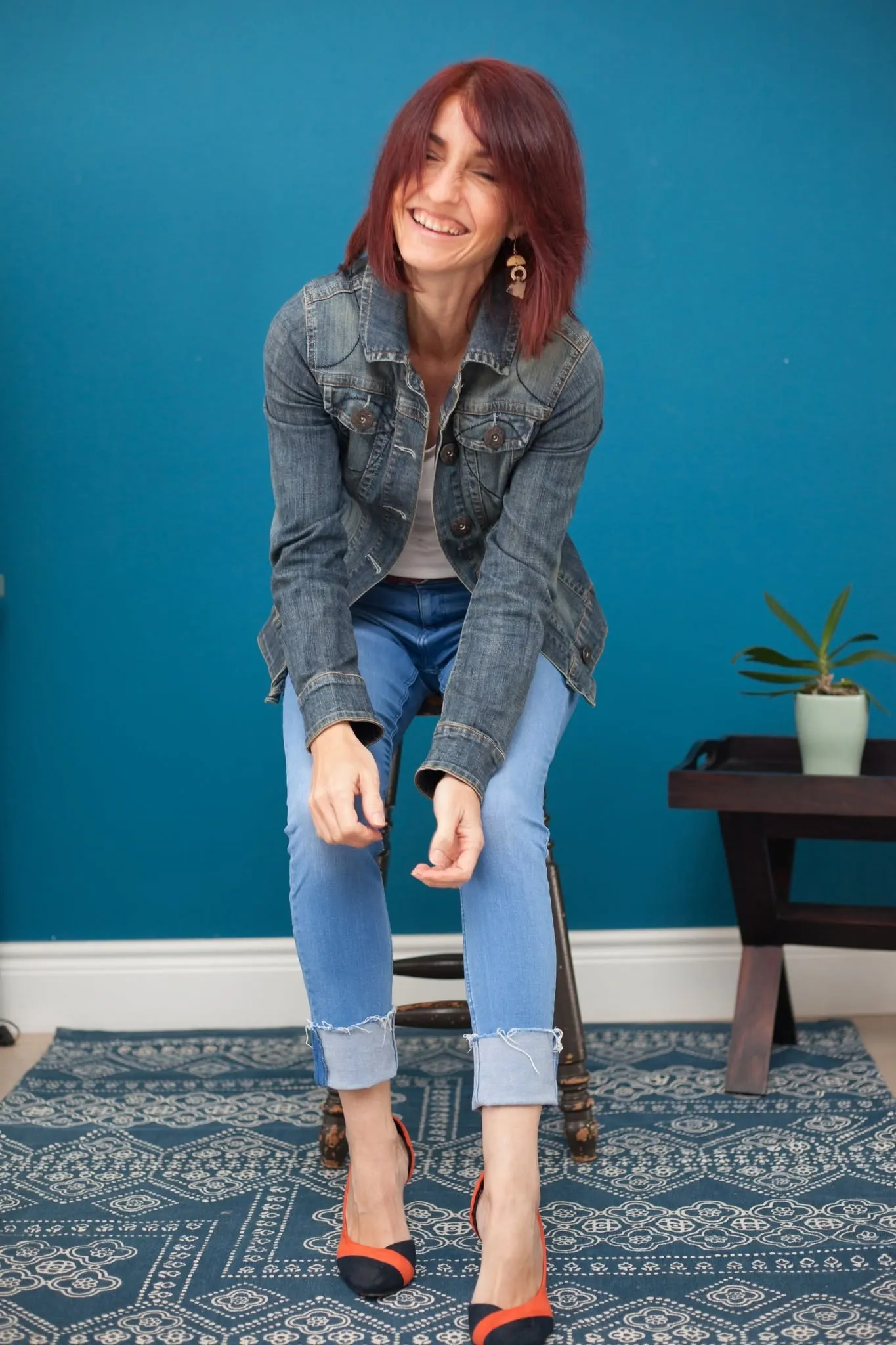 Full-length personal branding portrait of a smiling woman seated on a stool in front of a teal wall