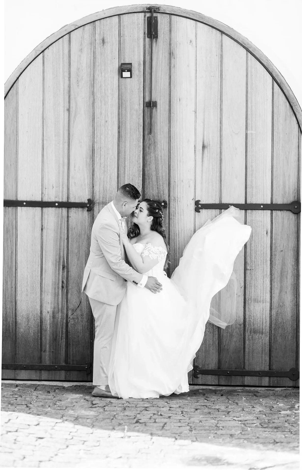 Black and white photo of a bride and groom nose to nose in front of arched wooden doors, her skirt lifted by the wind.