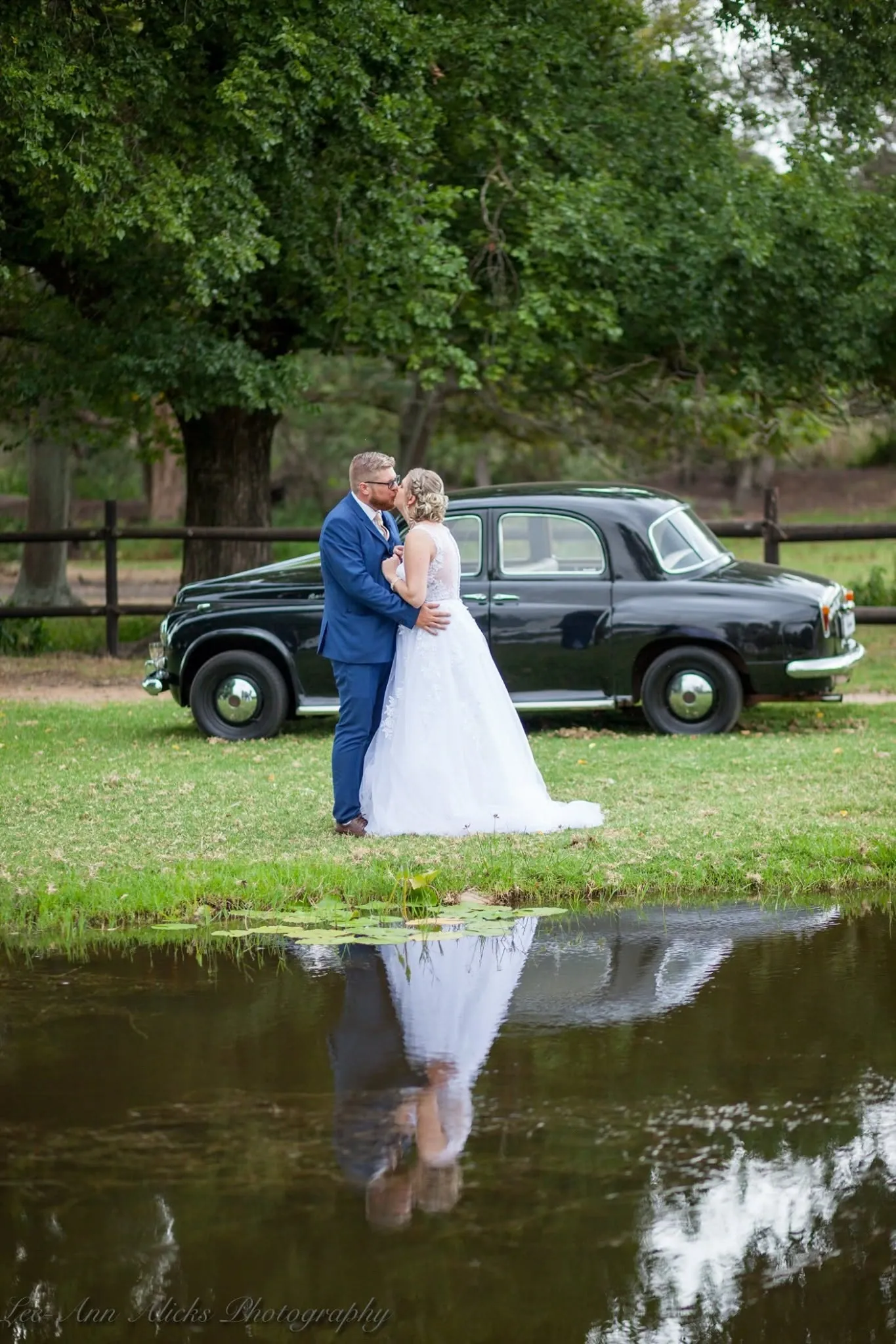 Bride and groom kissing beside a black vintage car, the whole scene reflected in a pond